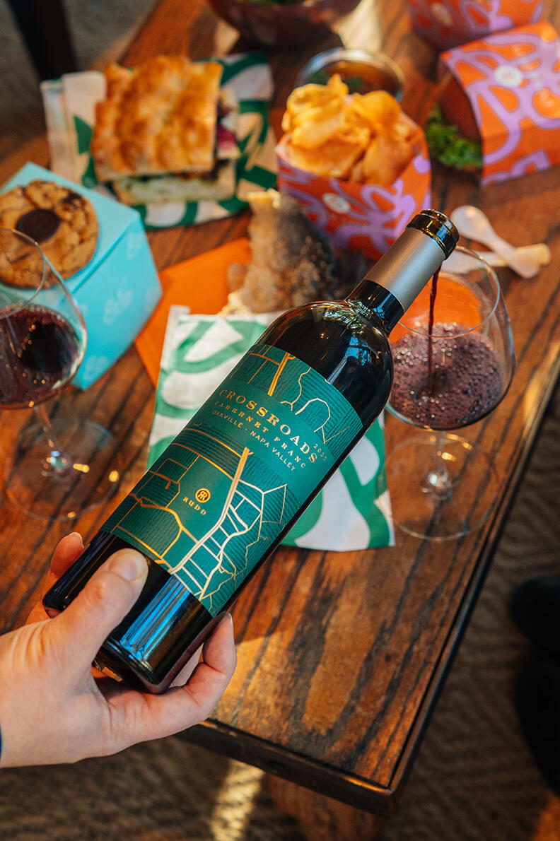 An afternoon pour of Crossroads Cabernet Franc anchors a vibrant scene with artisanal sandwiches scattered across a rustic wooden table. A moment to enjoy the food and wine together among friends.