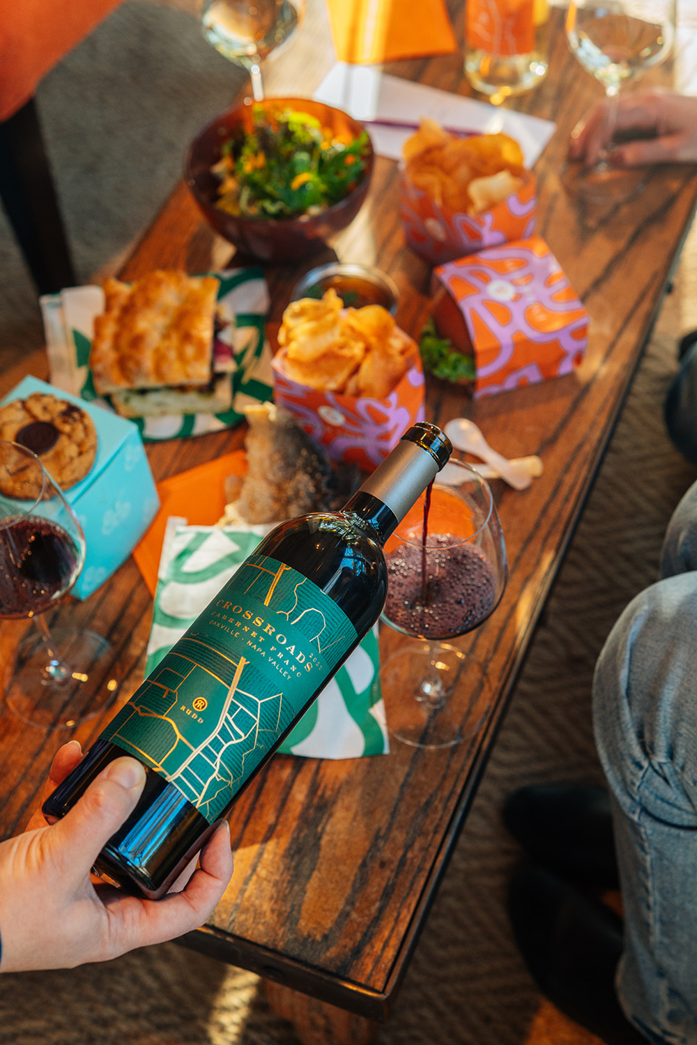 An afternoon pour of Crossroads Cabernet Franc anchors a vibrant scene with artisanal sandwiches scattered across a rustic wooden table. A moment to enjoy the food and wine together among friends.